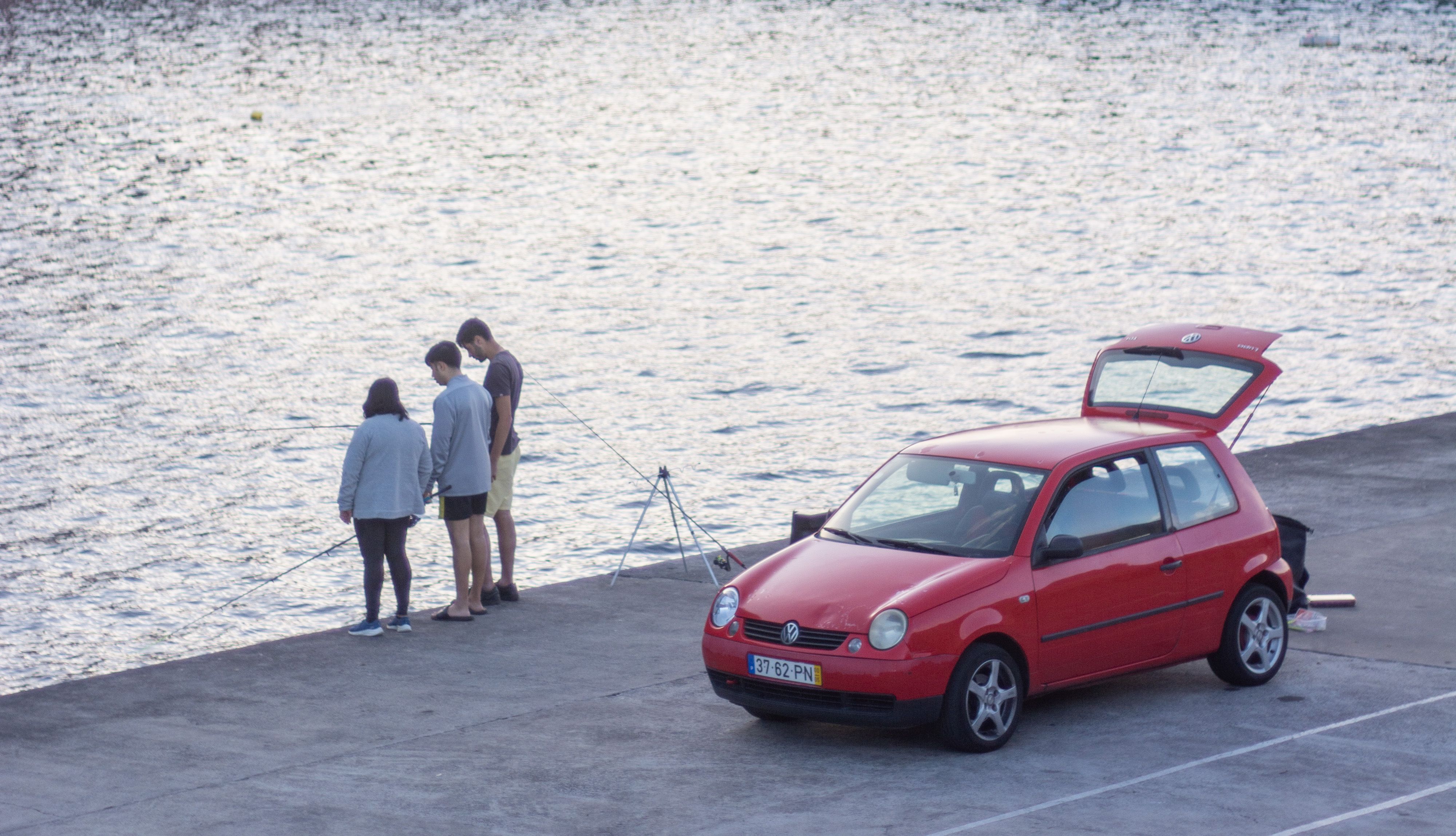 Three people standing in the harbor fishing in front of a red car