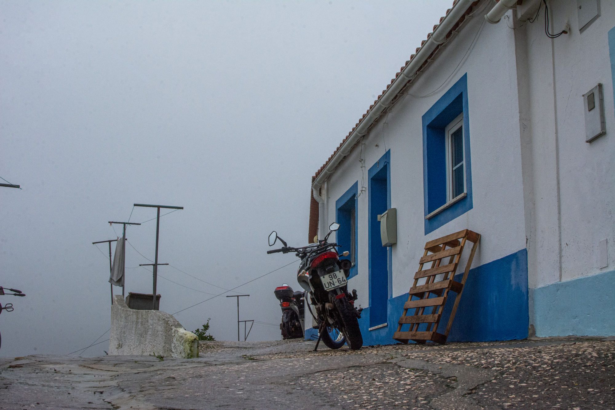 A parked motorcycle by a blue and white building on a cobblestone street in a misty and overcast setting.
