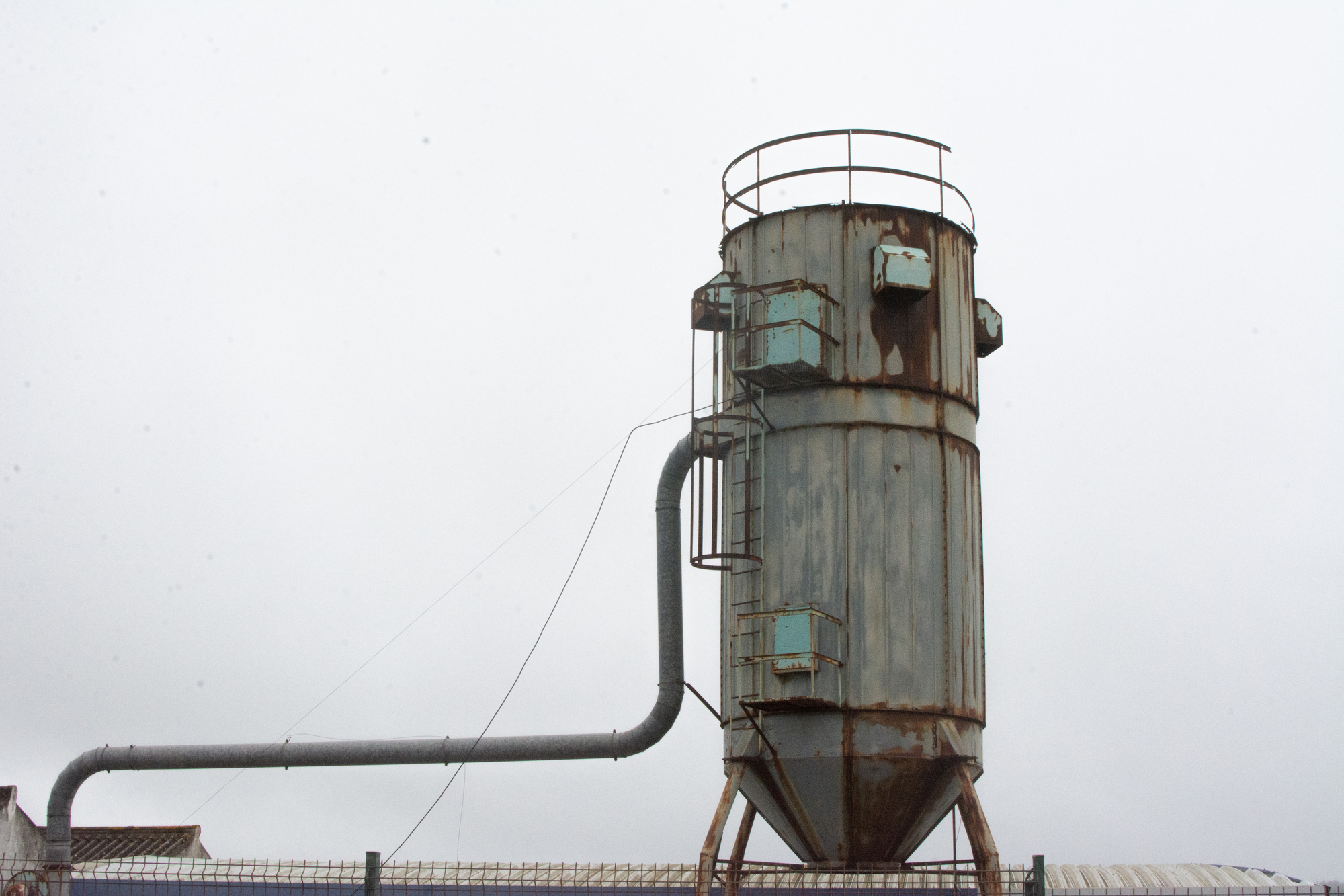 Rusty industrial silo with pipe against a cloudy sky.